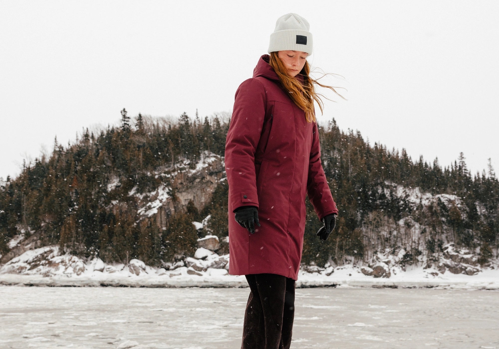 Femme dans un paysage hivernal portant un manteau d'hiver mi-long Audvik bordeaux et une tuque en tricot beige. 