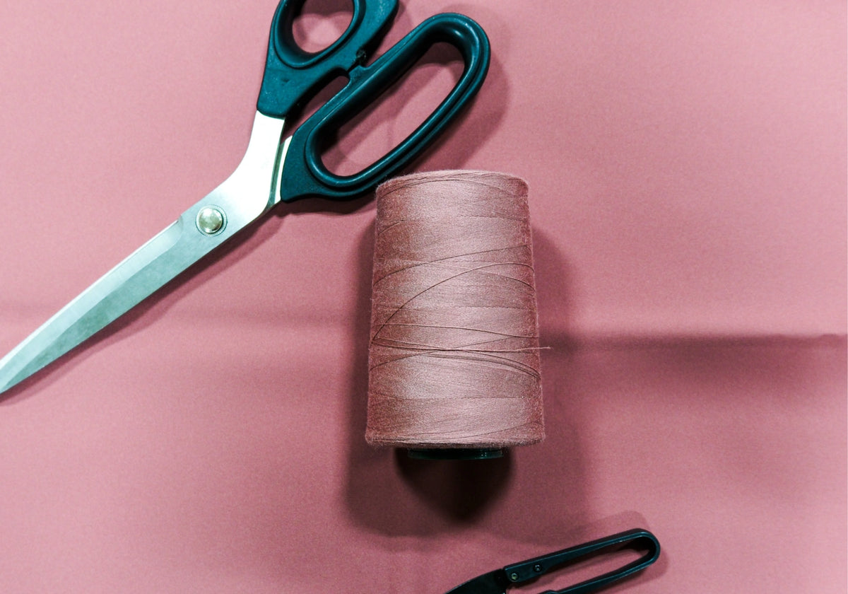 Photo d'une bobine de fil et d'une paire de ciseaux posés sur un tissu rose utilisé pour la création de manteaux d'hiver Audvik. 